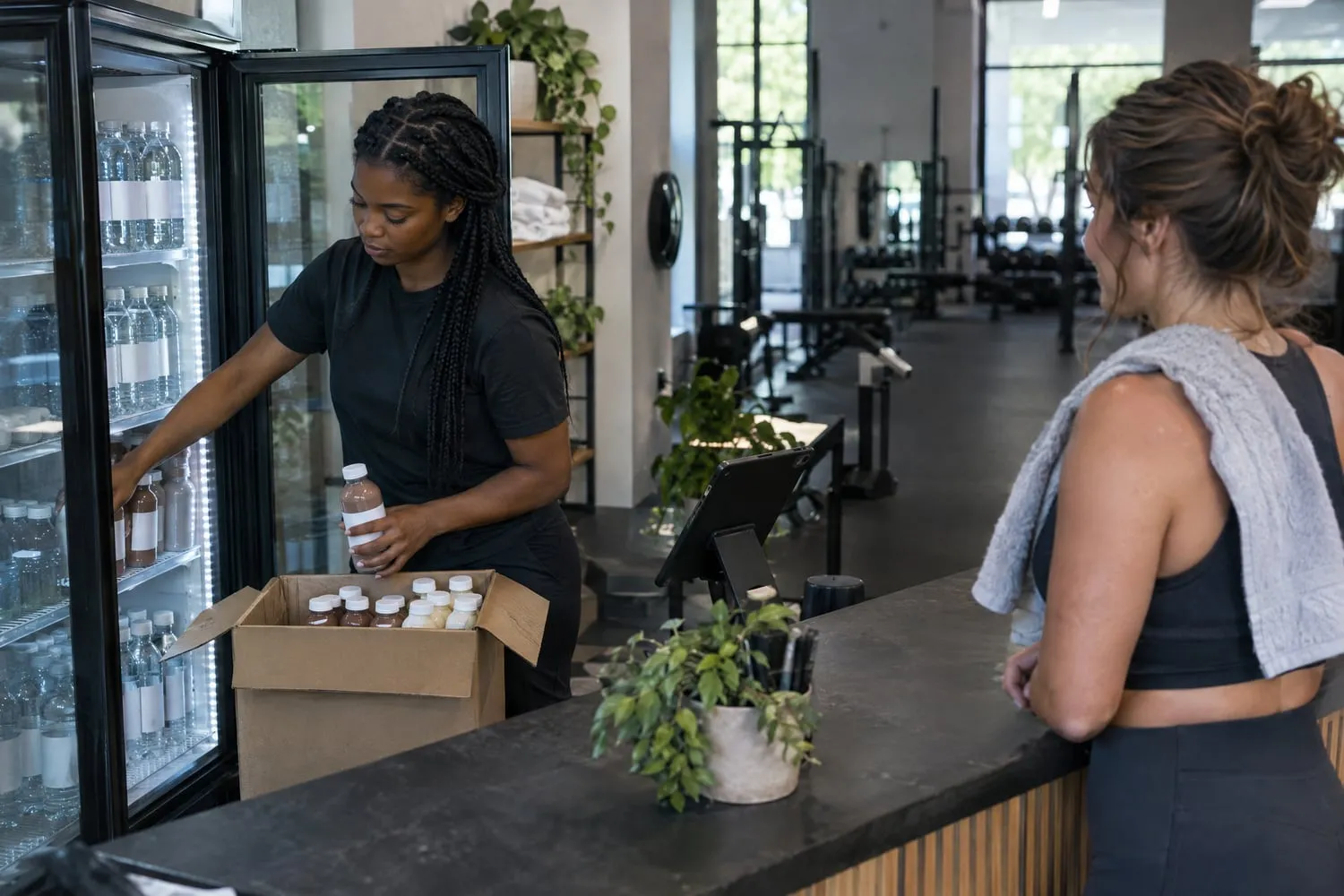 A front desk staffer stocks a glass-door cooler with water and protein shakes behind a gym's counter as a member waits